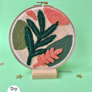 Peut inclure: Un kit de broderie au poinçon DIY avec un motif abstrait coloré composé de feuilles vertes et de formes de couleur pêche. Le motif est réalisé sur un cercle de broderie en bois et présenté sur un support en bois. Le texte "DIY Kit" est visible dans le coin inférieur gauche de l'image.