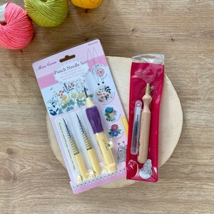 May include: A set of three punch needle tools in a clear plastic package with a floral design. The tools are yellow and purple with a spring mechanism. The package also includes a small wooden tool in a red plastic bag. The tools are on a wooden surface with three balls of yarn in the background.