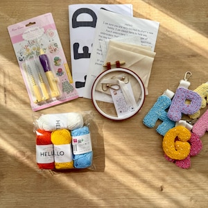May include: A craft kit containing punch needles, an embroidery hoop, yarn, and instructions. The yarn is in white, red, yellow, and blue. The kit includes the text "Hello" and "FD".
