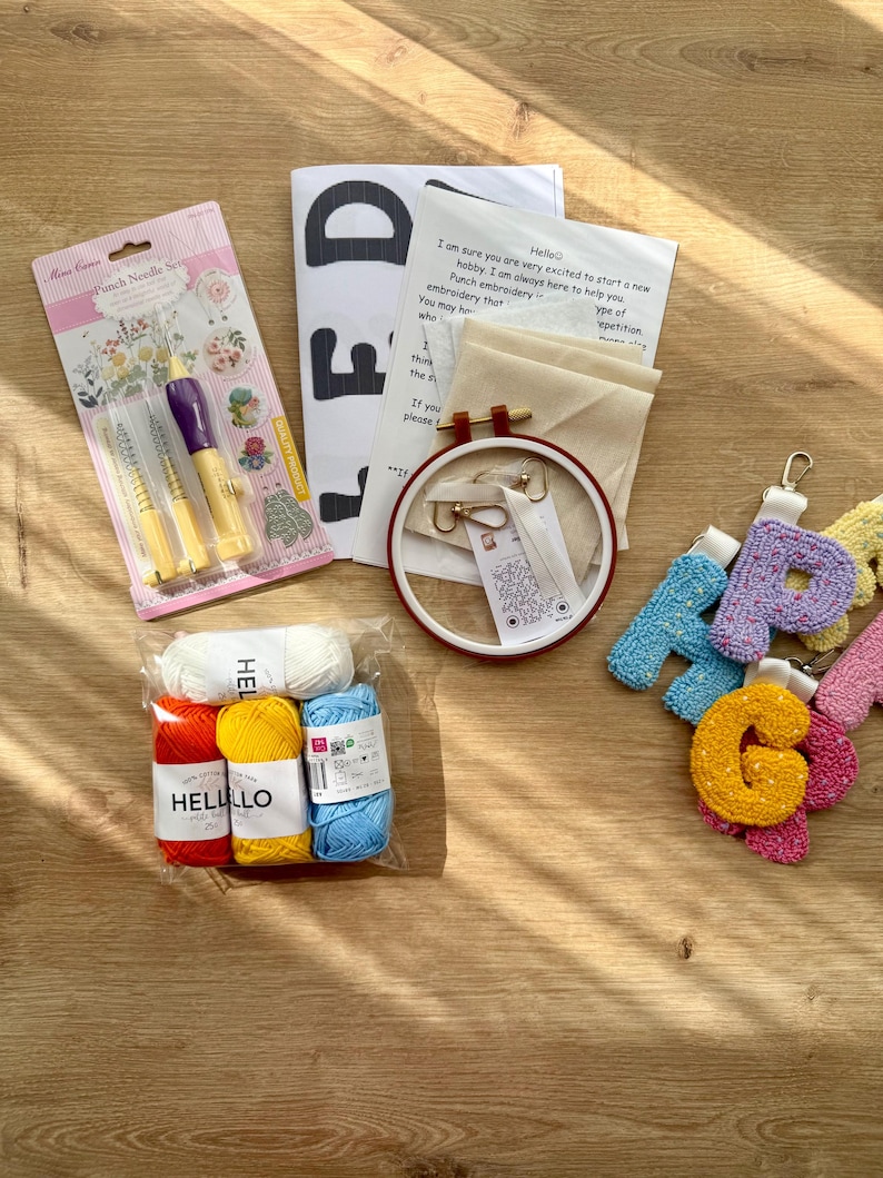 May include: A craft kit containing punch needles, an embroidery hoop, yarn, and instructions. The yarn is in white, red, yellow, and blue. The kit includes the text "Hello" and "FD".