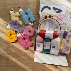 May include: A selection of colourful, tufted letter keyrings in yellow, blue, purple, pink, and orange, with silver-tone hardware. A craft kit includes yarn, a punch needle, embroidery hoop, and other supplies, all arranged on a wooden surface.