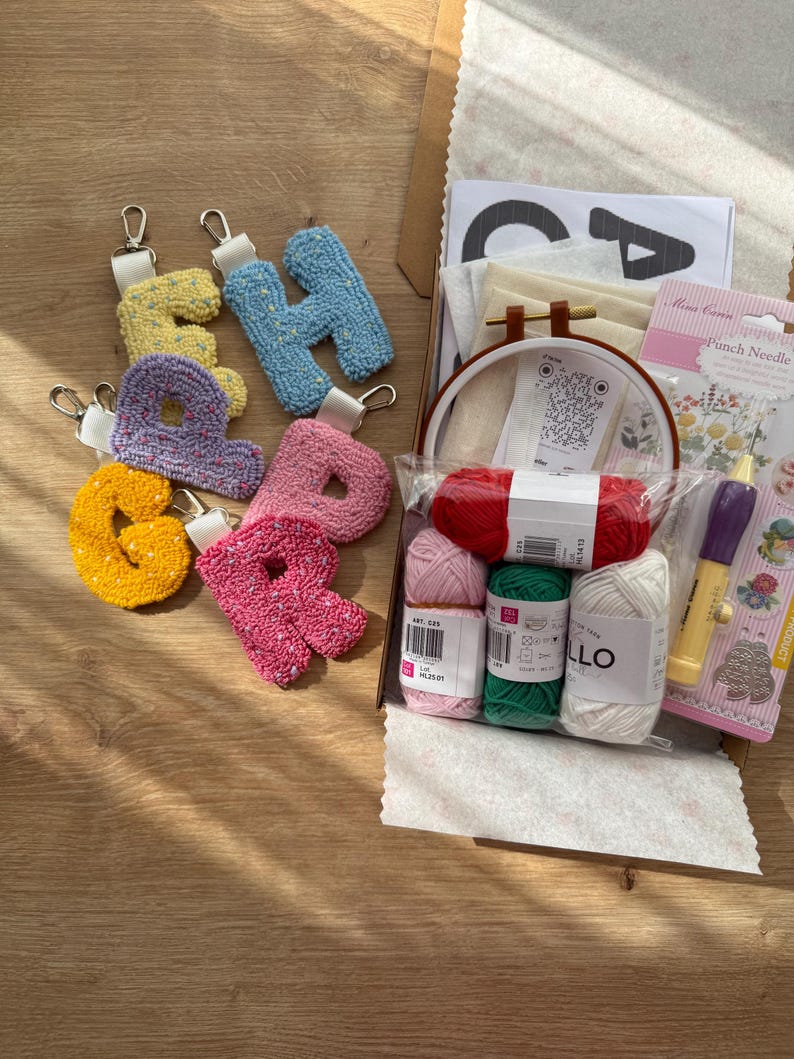 May include: A selection of colourful, tufted letter keyrings in yellow, blue, purple, pink, and orange, with silver-tone hardware. A craft kit includes yarn, a punch needle, embroidery hoop, and other supplies, all arranged on a wooden surface.