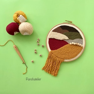 Op de afbeelding: Een kleurrijk abstract wandkleed met naaldvilt en een franje rand. Het kleed is gemaakt van verschillende gekleurde garens en houten kralen. Het kleed ligt op een groene achtergrond met garenbollen en een naaldviltnaald.