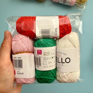 May include: A clear plastic package containing six small balls of yarn in various colours: red, pink, green, and off-white. Each ball has a label with the word "HELLO" and other text. The yarn is cotton.
