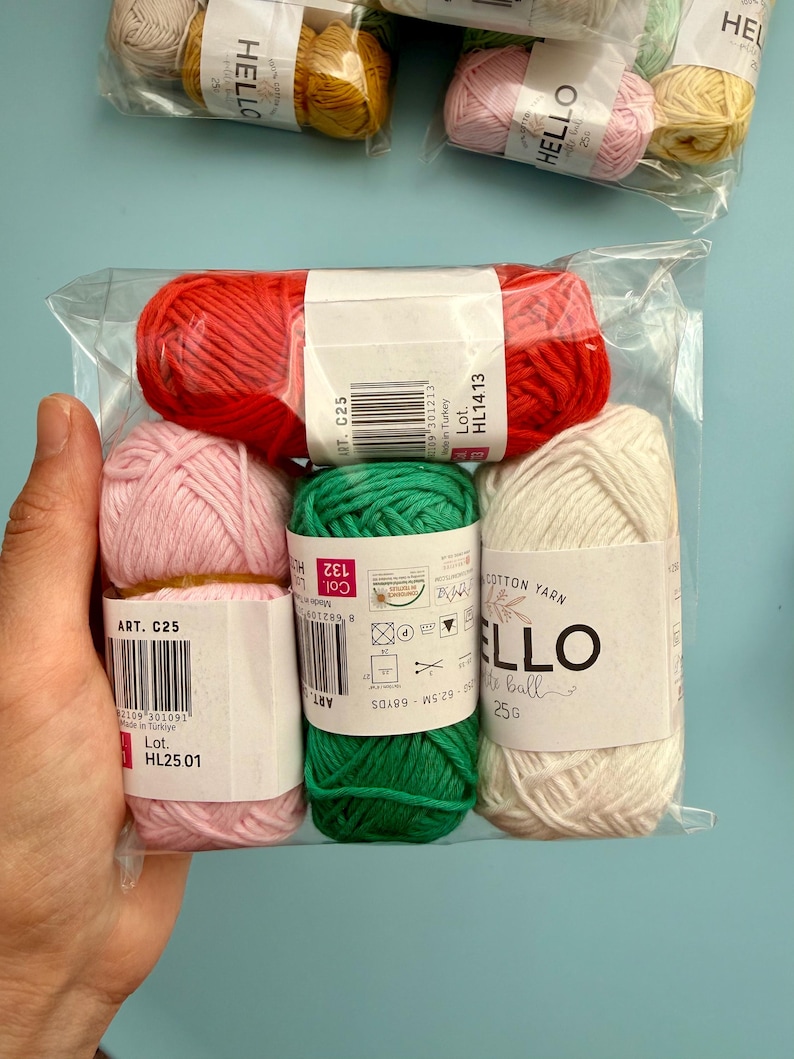 May include: A clear plastic package containing six small balls of yarn in various colours: red, pink, green, and off-white. Each ball has a label with the word "HELLO" and other text. The yarn is cotton.