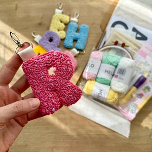 May include: A pink letter "R" keyring with a white strap and silver clasp. Other colourful letter keyrings are visible in the background, along with craft supplies including yarn and a small embroidery hoop. The letters appear to be made with a punch needle technique.