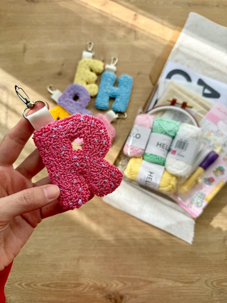 May include: A pink letter "R" keyring with a white strap and silver clasp. Other colourful letter keyrings are visible in the background, along with craft supplies including yarn and a small embroidery hoop. The letters appear to be made with a punch needle technique.