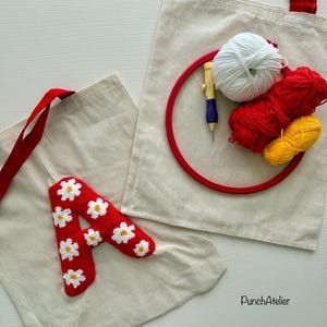 Op de afbeelding: Twee beige draagtassen met rode handvatten. Eén tas heeft een rode, met madeliefjes bedekte letter "A". De andere tas bevat een rode borduurring, witte, rode en gele wol en een punch needle tool. De tekst "PunchAtelier" is zichtbaar.