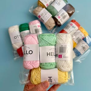 May include: A selection of multi-coloured yarn balls, including pink, green, yellow, and white, packaged in clear plastic. Each ball is labelled with the text "HELLO" or "HEL" and the weight of 25g. The yarn is displayed against a light blue background.