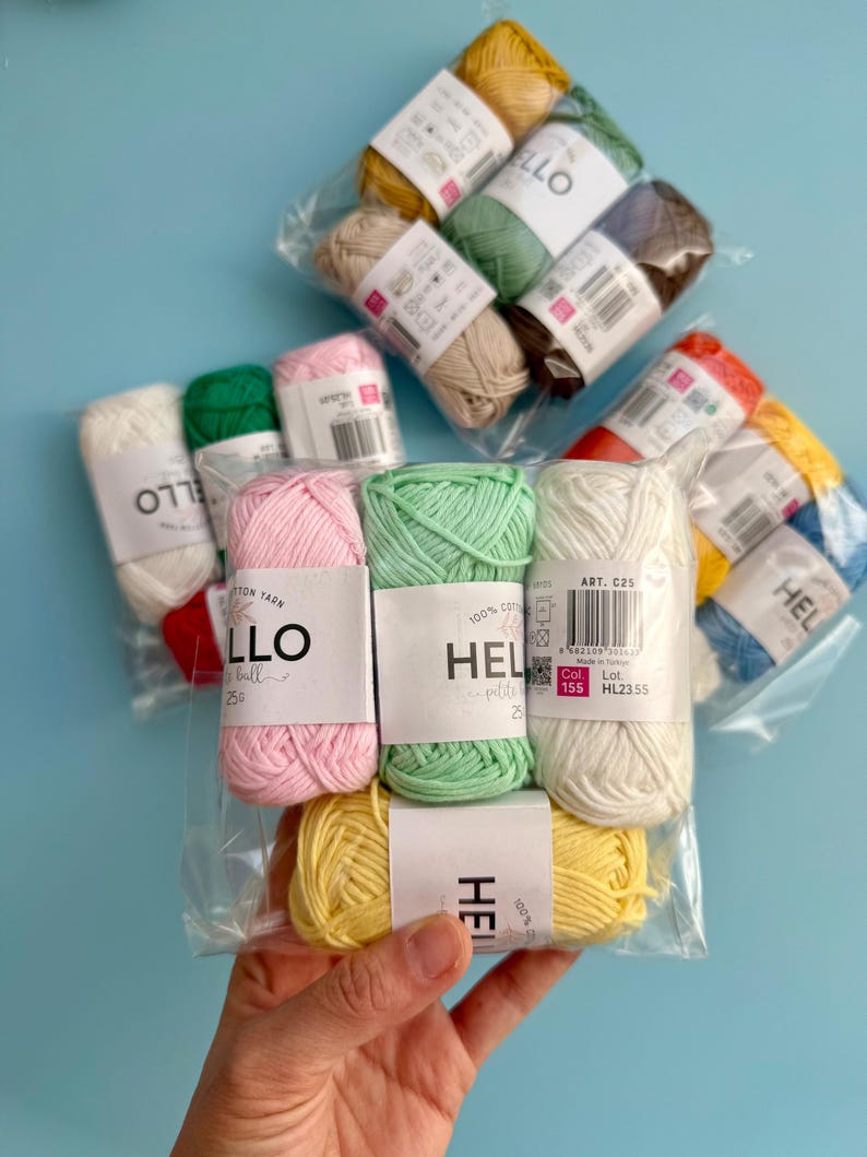 May include: A selection of multi-coloured yarn balls, including pink, green, yellow, and white, packaged in clear plastic. Each ball is labelled with the text "HELLO" or "HEL" and the weight of 25g. The yarn is displayed against a light blue background.