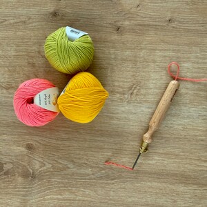 May include: Three balls of yarn in pink, yellow, and green colors, along with a wooden punch needle tool and a length of red thread on a wooden surface. The yarn balls have labels with the text "50% Acrylic 50% Cotton" on them.