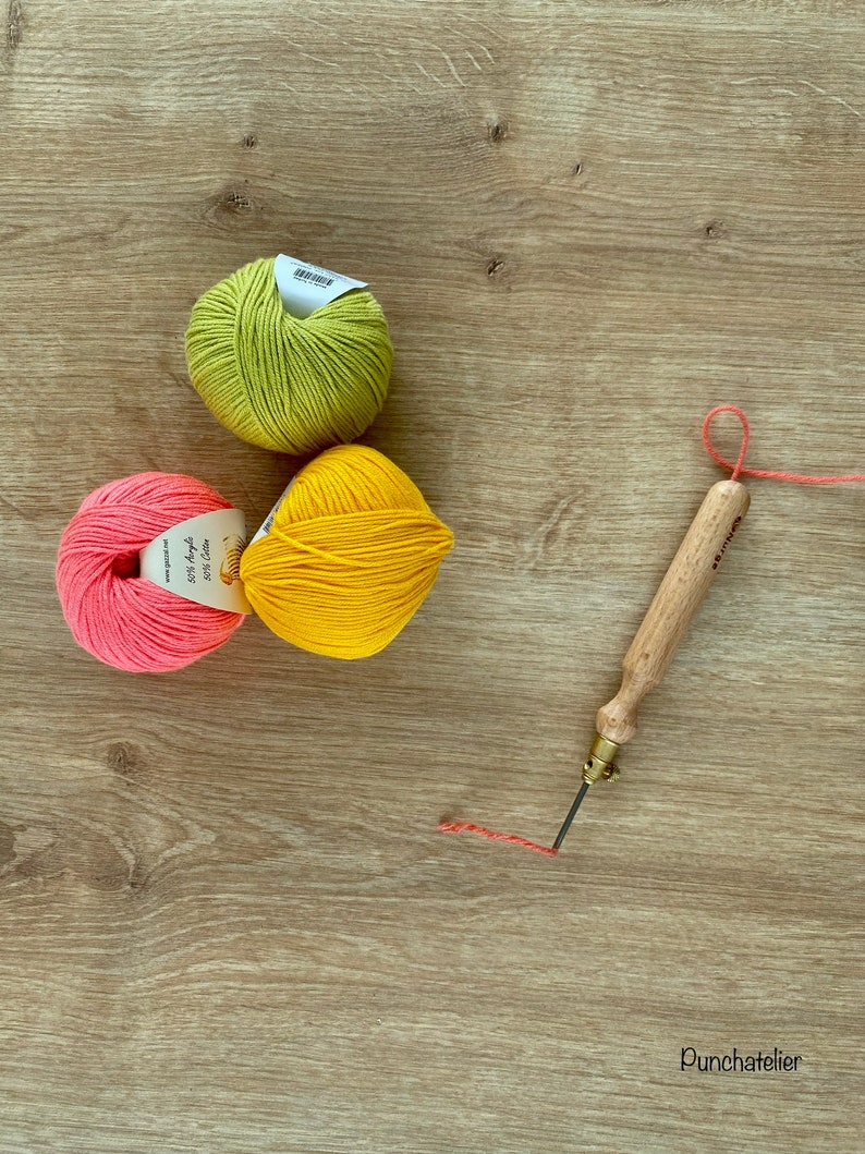 May include: Three balls of yarn in pink, yellow, and green colors, along with a wooden punch needle tool and a length of red thread on a wooden surface. The yarn balls have labels with the text "50% Acrylic 50% Cotton" on them.