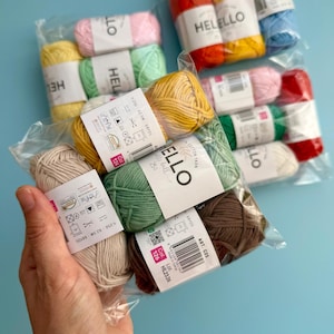 May include: A selection of multi-coloured cotton yarn skeins in clear plastic packaging. The yarn is available in various colours, including yellow, pink, green, and brown. Each skein is labelled with the brand name "HELLELLO" and product information.