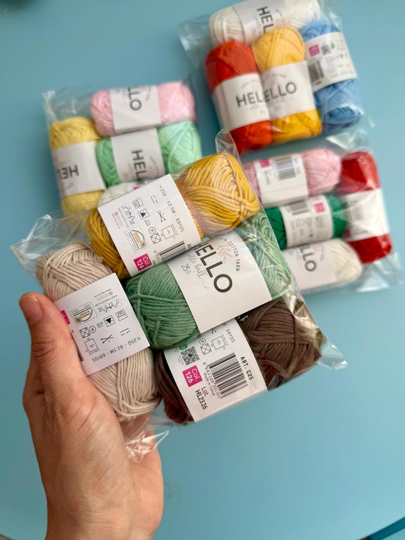May include: A selection of multi-coloured cotton yarn skeins in clear plastic packaging. The yarn is available in various colours, including yellow, pink, green, and brown. Each skein is labelled with the brand name "HELLELLO" and product information.