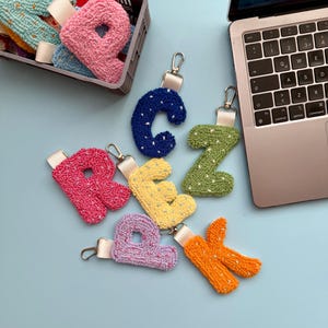 May include: A selection of colourful, handmade letter keyrings. The letters are in various colours, including pink, blue, green, yellow, purple, and orange. Each keyring has a silver clasp and a white ribbon. Some letters are in a grey basket.