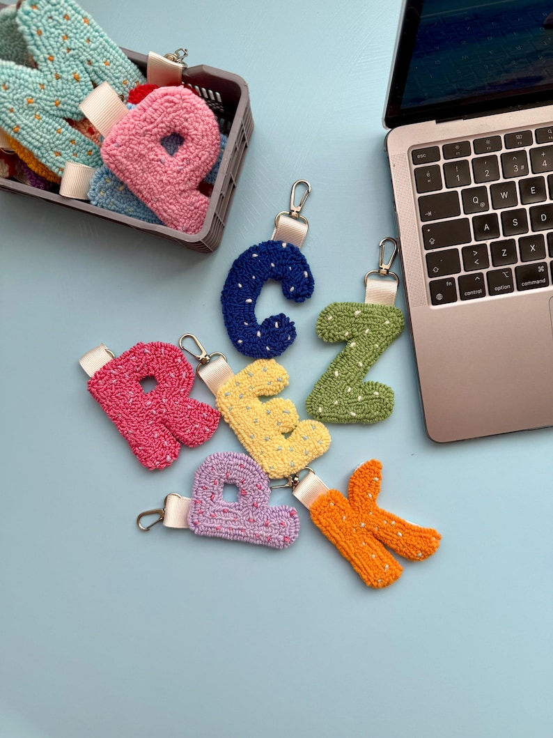 May include: A selection of colourful, handmade letter keyrings. The letters are in various colours, including pink, blue, green, yellow, purple, and orange. Each keyring has a silver clasp and a white ribbon. Some letters are in a grey basket.