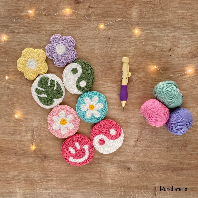 May include: A collection of eight colorful punch needle embroidery hoops with various designs, including flowers, a leaf, a yin yang symbol, and a smiley face. The hoops are arranged on a wooden surface with a punch needle and yarn balls.