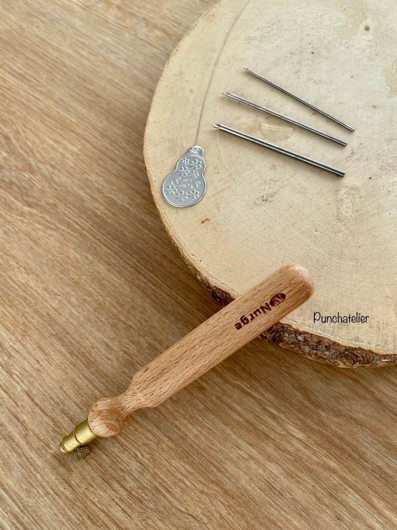 May include: A wooden punch needle with a brass tip and a wooden handle. The handle is engraved with the words "Punch Needle" and "Punchatelier".