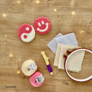 May include: Two pink and white yarn circles, one with a yin yang symbol and the other with a smiley face, are on a wooden surface with a needle tool, two balls of yarn, and a hoop with fabric inside.