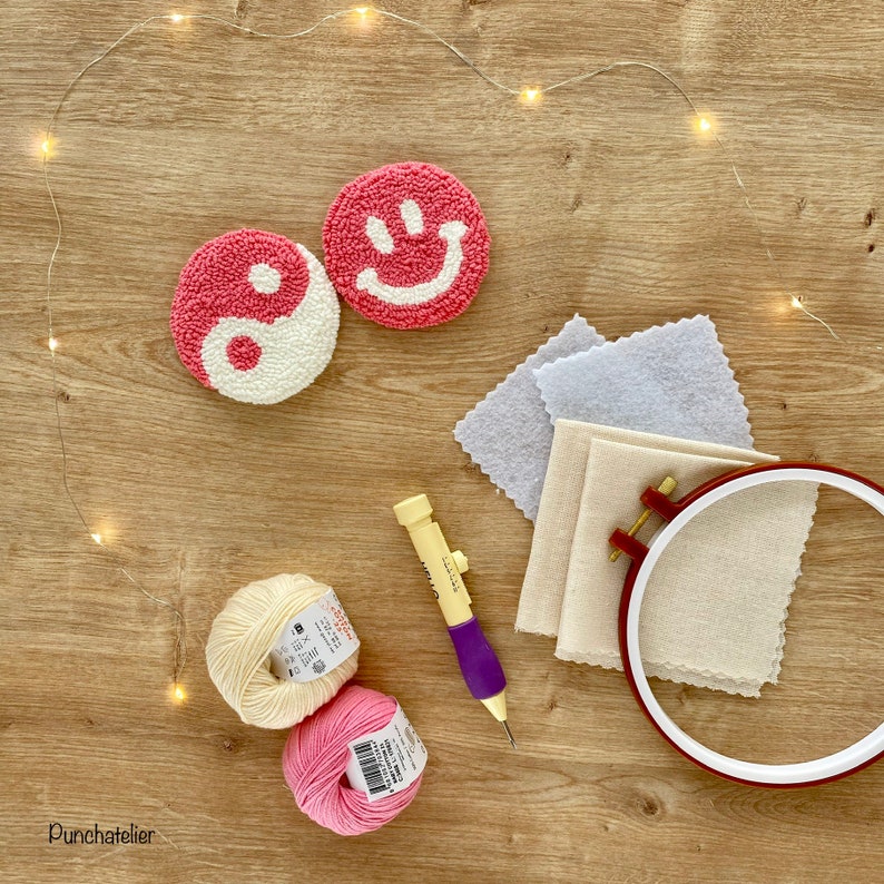 May include: Two pink and white yarn circles, one with a yin yang symbol and the other with a smiley face, are on a wooden surface with a needle tool, two balls of yarn, and a hoop with fabric inside.