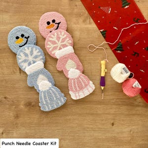 May include: Handmade punch needle coasters in the shape of a snowman, mittens, and a hat. The coasters are in blue and pink, with white details. A punch needle tool, yarn, and a red fabric with a Christmas tree pattern are also visible. The text "Punch Needle Coaster Kit" is at the bottom.