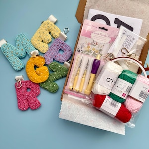 May include: A craft kit featuring colourful, textured letter keychains and punch needle supplies. The keychains are in shades of blue, yellow, purple, orange, and green. The kit includes yarn, needles, and a hoop, on a light blue surface.