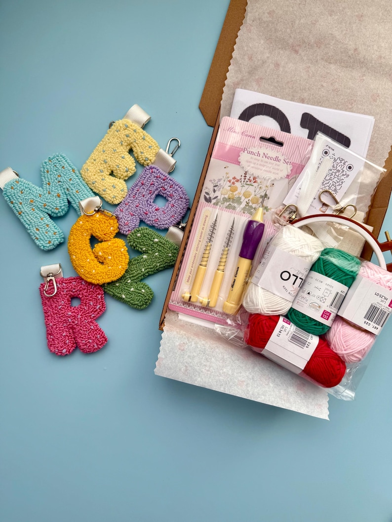 May include: A craft kit featuring colourful, textured letter keychains and punch needle supplies. The keychains are in shades of blue, yellow, purple, orange, and green. The kit includes yarn, needles, and a hoop, on a light blue surface.