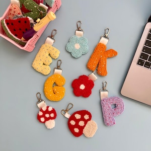 May include: A selection of handmade keyrings in various designs. The keyrings feature the letters E, G, K, and P, plus flower and mushroom shapes. Each keyring has a metal clasp and a white ribbon. The keyrings are made from a textured, looped fabric.
