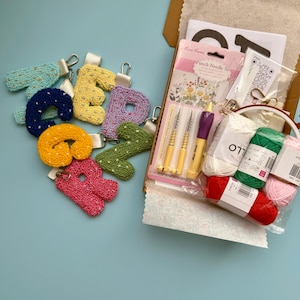 May include: A selection of embroidered letter keyrings in shades of yellow, pink, green, and blue are arranged on a light blue surface. A craft kit containing punch needles and yarn is open in a cardboard box. The kit includes various colours of yarn.