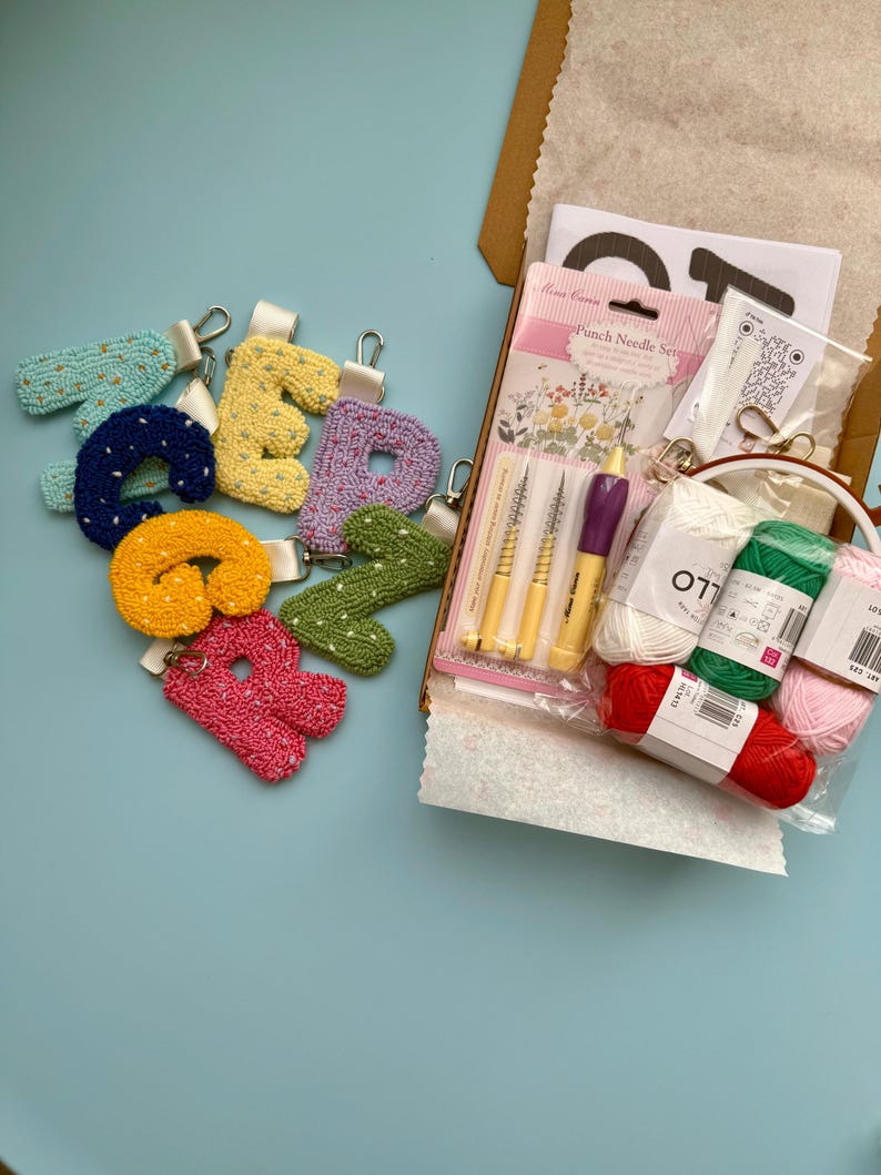 May include: A selection of embroidered letter keyrings in shades of yellow, pink, green, and blue are arranged on a light blue surface. A craft kit containing punch needles and yarn is open in a cardboard box. The kit includes various colours of yarn.