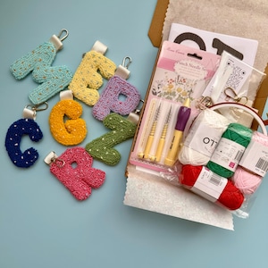 May include: A selection of embroidered letter keyrings in shades of blue, yellow, and pink. A craft kit with punch needles and yarn in red, green, and pink is also visible, along with a paper bag, all on a light blue surface.