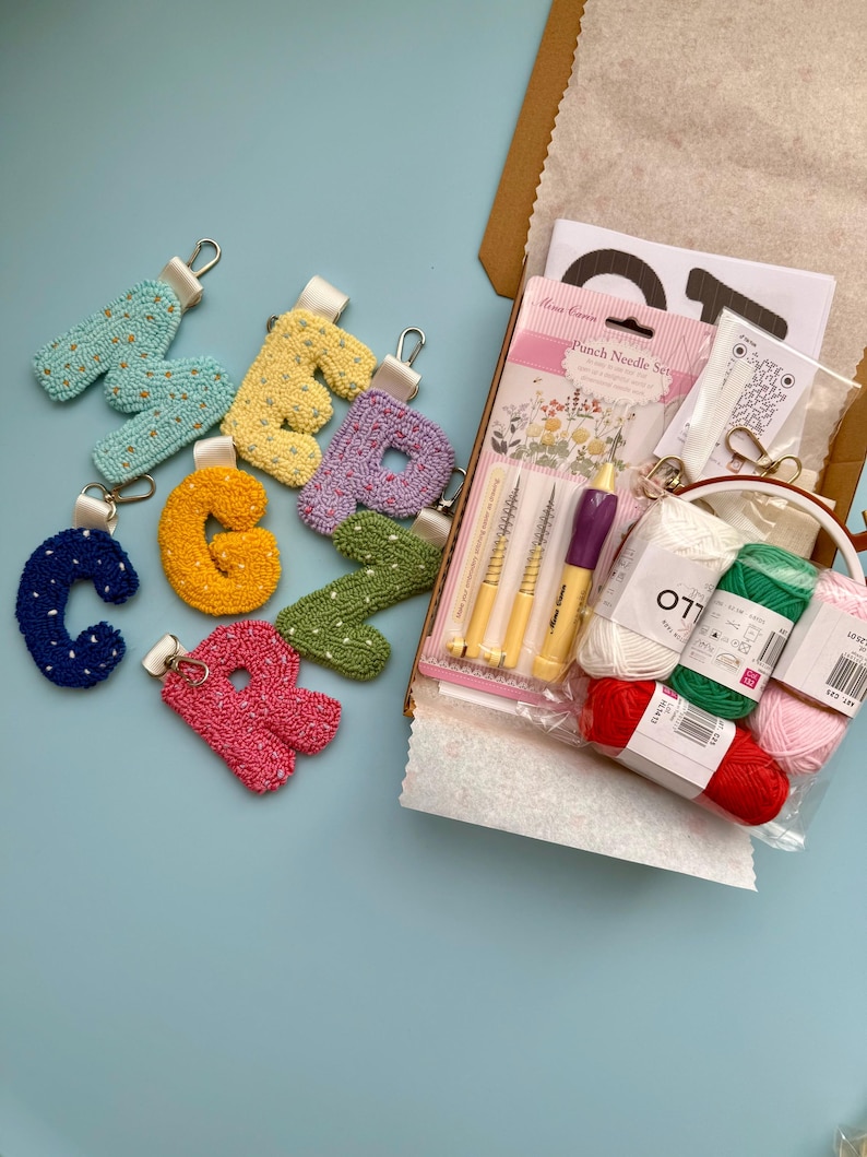 May include: A selection of embroidered letter keyrings in shades of blue, yellow, and pink. A craft kit with punch needles and yarn in red, green, and pink is also visible, along with a paper bag, all on a light blue surface.