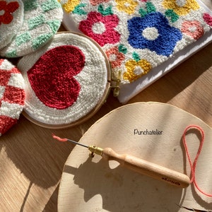 May include: A close-up of a punch needle tool with a wooden handle and a metal needle. The tool is on a wooden surface with a red yarn and a white hoop with a red heart design. There are other hoops with colorful floral designs in the background.