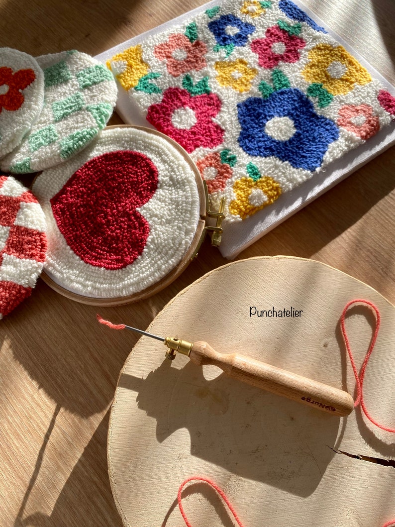 May include: A close-up of a punch needle tool with a wooden handle and a metal needle. The tool is on a wooden surface with a red yarn and a white hoop with a red heart design. There are other hoops with colorful floral designs in the background.