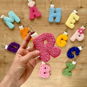 Può includere: Una collezione di portachiavi a forma di lettera realizzati con tecnica tufting in vari colori, tra cui rosa, blu, giallo, arancione, viola e verde. Ogni lettera presenta un nastro bianco con un moschettone in metallo argentato. La lettera R è in primo piano.