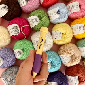 May include: A close-up of a purple and yellow crochet hook with a variety of colorful yarn balls in the background. The yarn balls are arranged in a circle around the crochet hook. The yarn balls are all different colors, including pink, red, green, blue, yellow, and brown.