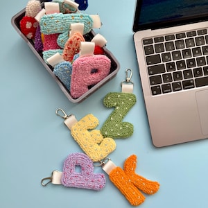 May include: A selection of handmade letter keyrings in various colours. The keyrings are made from textured fabric and include shades of pink, yellow, green, and orange. Some are in a container, others are displayed on a light blue surface.