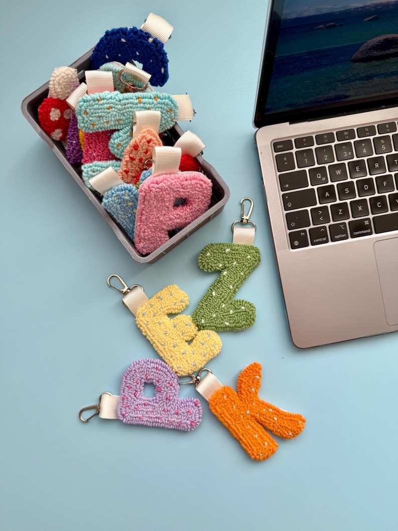 May include: A selection of handmade letter keyrings in various colours. The keyrings are made from textured fabric and include shades of pink, yellow, green, and orange. Some are in a container, others are displayed on a light blue surface.