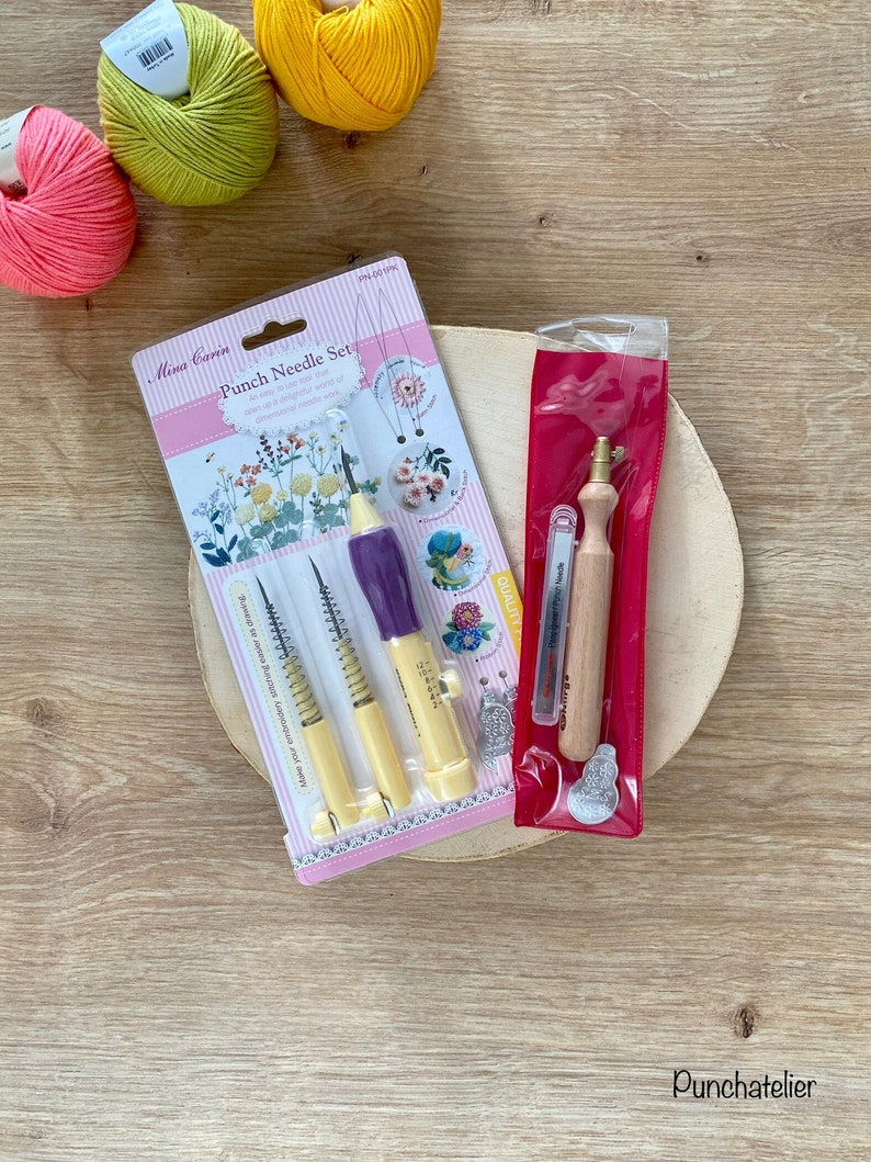 May include: A set of three punch needle tools in a clear plastic package with a floral design. The tools are yellow and purple with a spring mechanism. The package also includes a small wooden tool in a red plastic bag. The tools are on a wooden surface with three balls of yarn in the background.