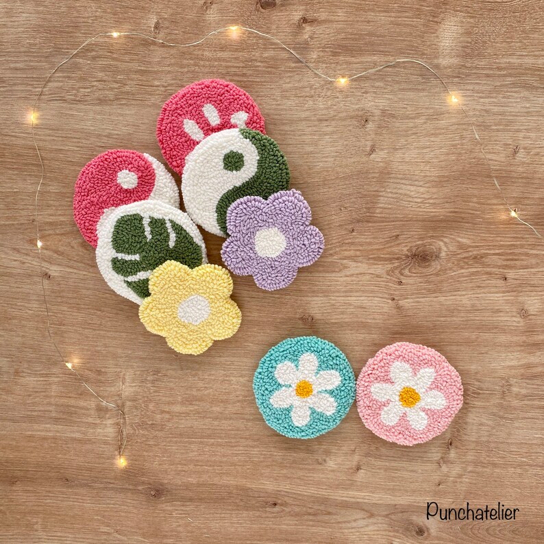 May include: A set of seven colorful, hand-stitched, round, wool coasters with various designs, including a flower, a leaf, a yin-yang symbol, and a hand. The coasters are arranged on a wooden surface with string lights.