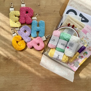 May include: A selection of colourful, textured letter keychains spelling "ERAH" in yellow, pink, blue, purple, and orange. The keychains are shown with a craft kit containing yarn, a hoop, and other supplies.