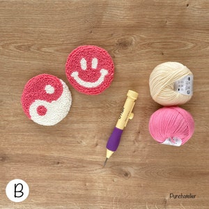 May include: Two pink and white yarn punch needle projects, one with a yin yang design and the other with a smiley face. A purple and yellow punch needle tool and two balls of yarn, one cream and one pink, are also included.