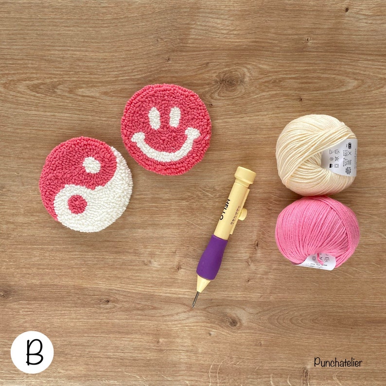 May include: Two pink and white yarn punch needle projects, one with a yin yang design and the other with a smiley face. A purple and yellow punch needle tool and two balls of yarn, one cream and one pink, are also included.