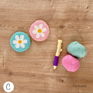 May include: Two punch needle projects, one in pink and one in blue, with a daisy design. The projects are on a wooden surface with a punch needle, pink and blue yarn, and the word "Punchatelier" written on the surface.
