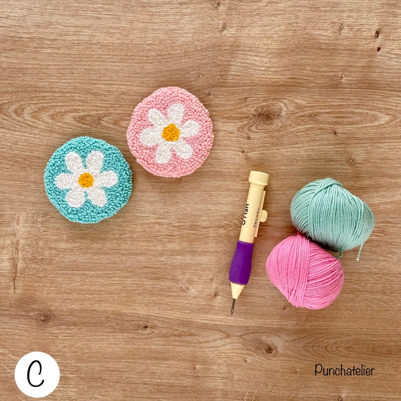 May include: Two punch needle projects, one in pink and one in blue, with a daisy design. The projects are on a wooden surface with a punch needle, pink and blue yarn, and the word "Punchatelier" written on the surface.