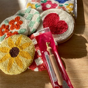 May include: A set of five colorful punch needle embroidery hoops with various designs, including a sunflower, a heart, and a checkered pattern. A punch needle tool is also included in a clear plastic pouch.