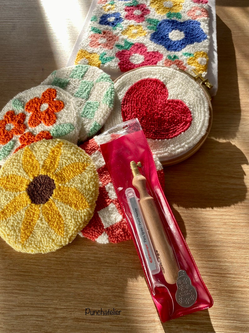 May include: A set of five colorful punch needle embroidery hoops with various designs, including a sunflower, a heart, and a checkered pattern. A punch needle tool is also included in a clear plastic pouch.