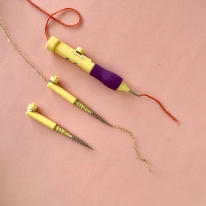 May include: Three punch needle tools on a pink background. Two are yellow with a spring and a needle. The third is purple and yellow with a needle and a knob to adjust the needle depth. A red thread is threaded through the purple tool and a gold thread is threaded through one of the yellow tools.