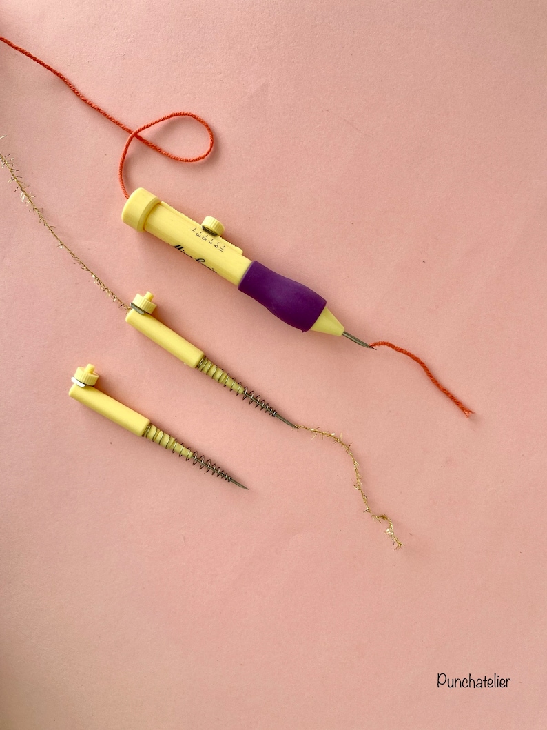 May include: Three punch needle tools on a pink background. Two are yellow with a spring and a needle. The third is purple and yellow with a needle and a knob to adjust the needle depth. A red thread is threaded through the purple tool and a gold thread is threaded through one of the yellow tools.