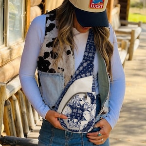 May include: A beige and navy blue baseball cap with a rectangular patch featuring a blue rabbit and the word LUCKY. A white long-sleeve shirt, a cow print vest with denim pockets, blue jeans, and a blue and white patterned crossbody sling bag.